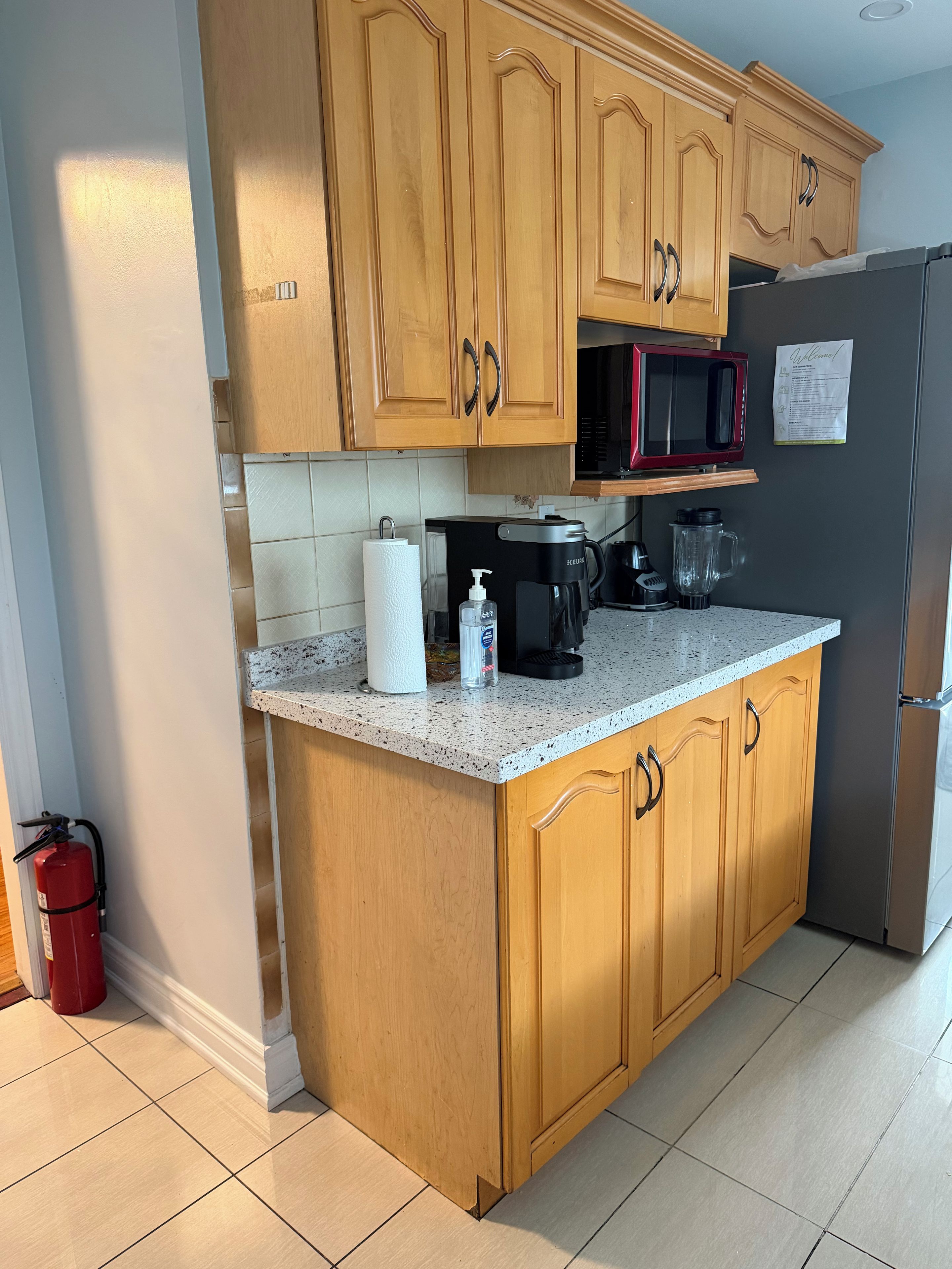 A kitchen with wooden cabinets, a stainless steel refrigerator, a microwave, a coffee maker, and a blender on the countertop.