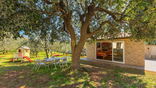 A tree-lined outdoor area with a play structure, a dining table with chairs, and a building with a glass door.