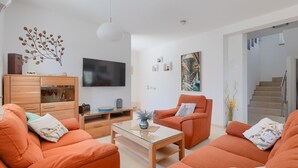 A modern living room with orange sofas, a glass coffee table, a wooden cabinet, a flat-screen TV, and a staircase.