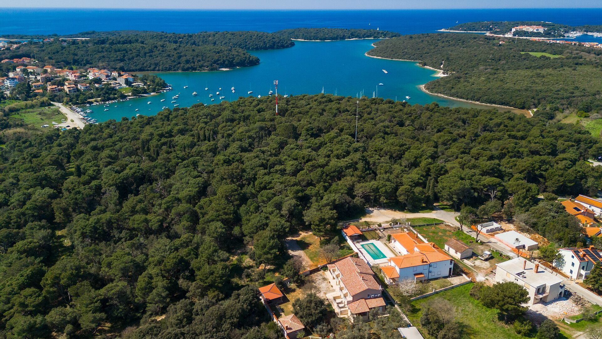 An aerial view of a coastal area with dense forest, a marina with boats, and residential buildings with red roofs.