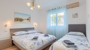 A double room with two beds, a chandelier, a beach-themed wall art, and a window with sheer curtains.