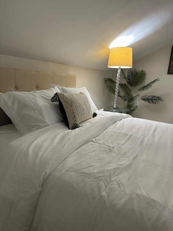 A neatly made bed with white linens and a beige headboard, a bedside lamp, and a potted plant. - Modern 3-bedroom house with WiFi and AC in North Rosedale Park (Detroit)