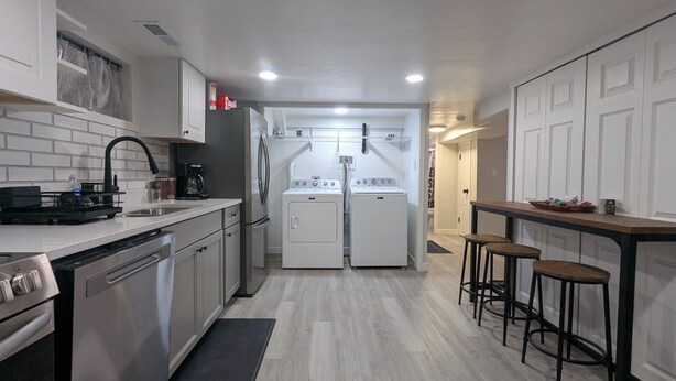A compact kitchen with a dishwasher, stove, and a built-in washer and dryer. There are stools at a small table and a black mat leading to the kitchen.