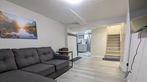 A modern living room with a grey sofa, a painting of a landscape, and a kitchen visible through an open doorway.