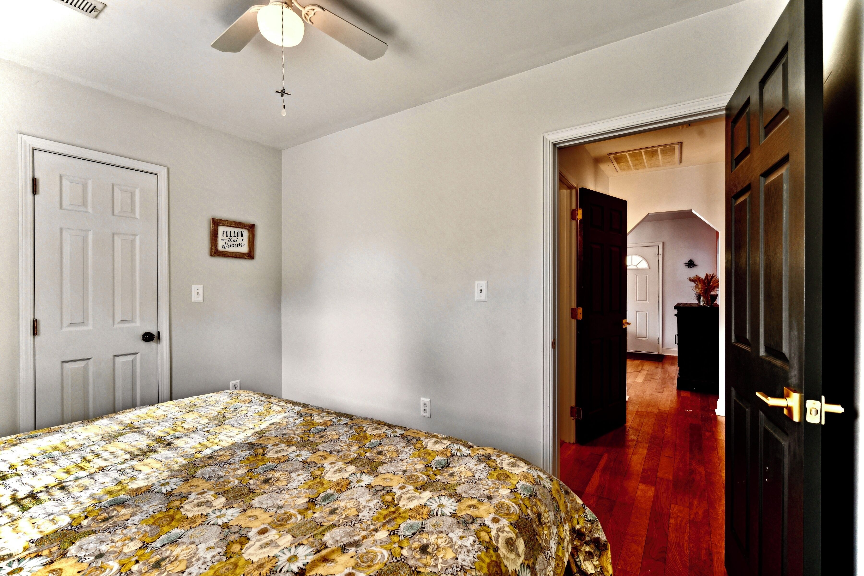 Un dormitorio con cubrecama floral, ventilador de techo, puerta blanca y pasillo que conduce a otra habitación.