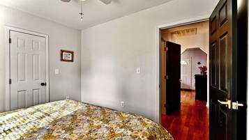 Un dormitorio con cubrecama floral, ventilador de techo, puerta blanca y pasillo que conduce a otra habitación.