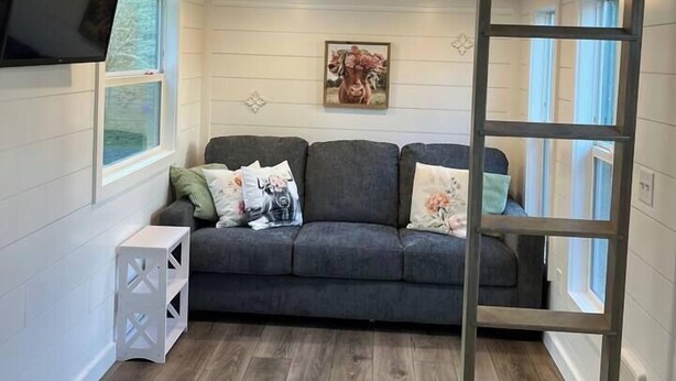 Ein Zimmer mit grauem Sofa, einer Hockerleiter, einem Fernseher und Holzwänden.