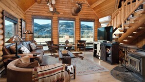 A log cabin interior with a fireplace, leather furniture, mounted animal heads, and large windows offering a mountain view. - SPACIOUS & Stunning 6-bedroom Log Cabin Home Great for Large Gatherings! (Emigrant)