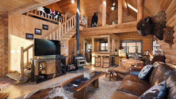 A log cabin interior with a wooden staircase, a fireplace, a television, a leather sofa, and a mounted bison head. - SPACIOUS & Stunning 6-bedroom Log Cabin Home Great for Large Gatherings! (Emigrant)