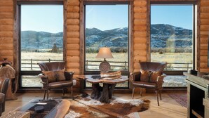 A log cabin interior with two leather chairs, a wooden table with a lamp, and large windows offering a view of snow-covered mountains. - SPACIOUS & Stunning 6-bedroom Log Cabin Home Great for Large Gatherings! (Emigrant)