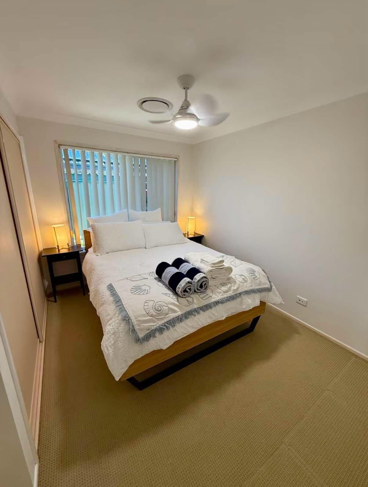 A neatly made bed with a white comforter and rolled towels, a ceiling fan, and a window with blinds.