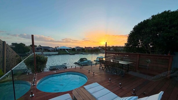 A wooden deck with a pool, lounge chairs, and a view of a body of water at sunset.