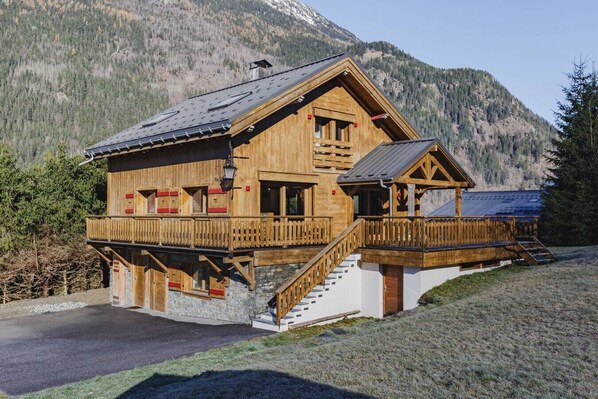 Un chalet de madera con balcón y una escalera que lleva al segundo piso, con montañas al fondo.
