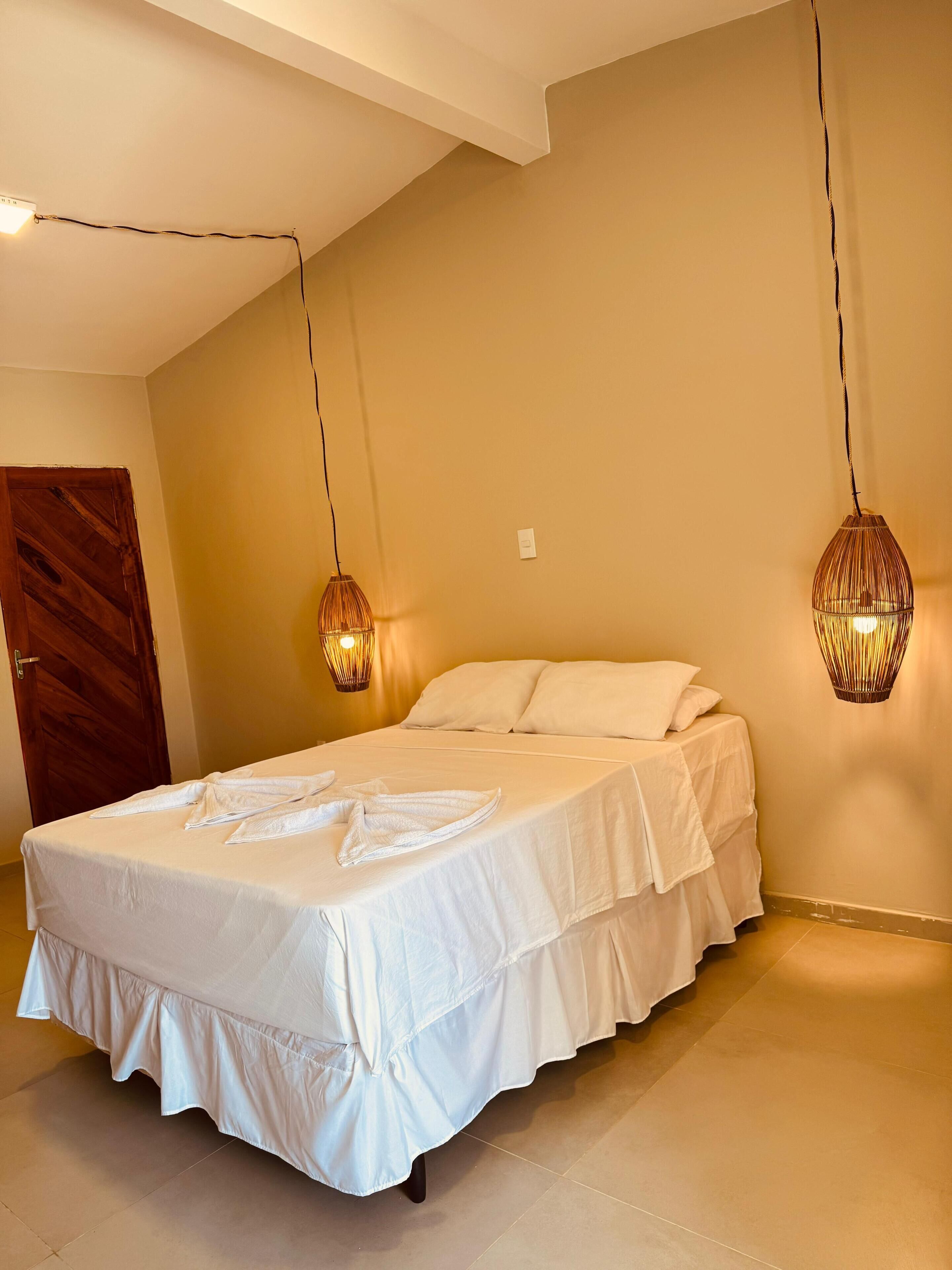 A bedroom with a bed, two hanging lamps, a wooden door, and a light switch on the wall.