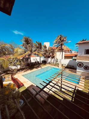 A swimming pool with a wooden deck, surrounded by palm trees and a white building with a tiled roof. - Pousada Villa Manzuá  (Cascavel)