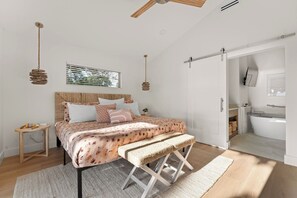 Un dormitorio con cama, mesita de noche, lámpara colgante y vista al baño.
