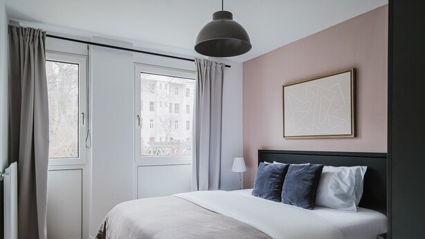 A bedroom with a bed, a bedside lamp, a window with curtains, and a framed picture on the wall.