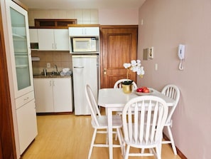 Une cuisine compacte avec des meubles blancs, un four à micro-ondes et un réfrigérateur. On y trouve une table à manger avec des chaises blanches et un vase rempli de fleurs.