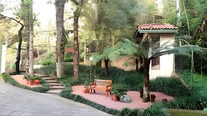 Un camino pavimentado que lleva a un banco, con un pequeño jardín, rodeado de árboles y un edificio con techo rojo.