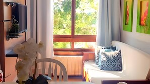 Una habitación con cama, una silla, una mesa con un florero y un televisor montado en la pared, además de una ventana con vistas a árboles.
