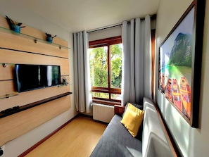 Een moderne woonkamer met een flatscreen televisie, een bank met kussens, een raam met gordijnen en een schilderij aan de muur.