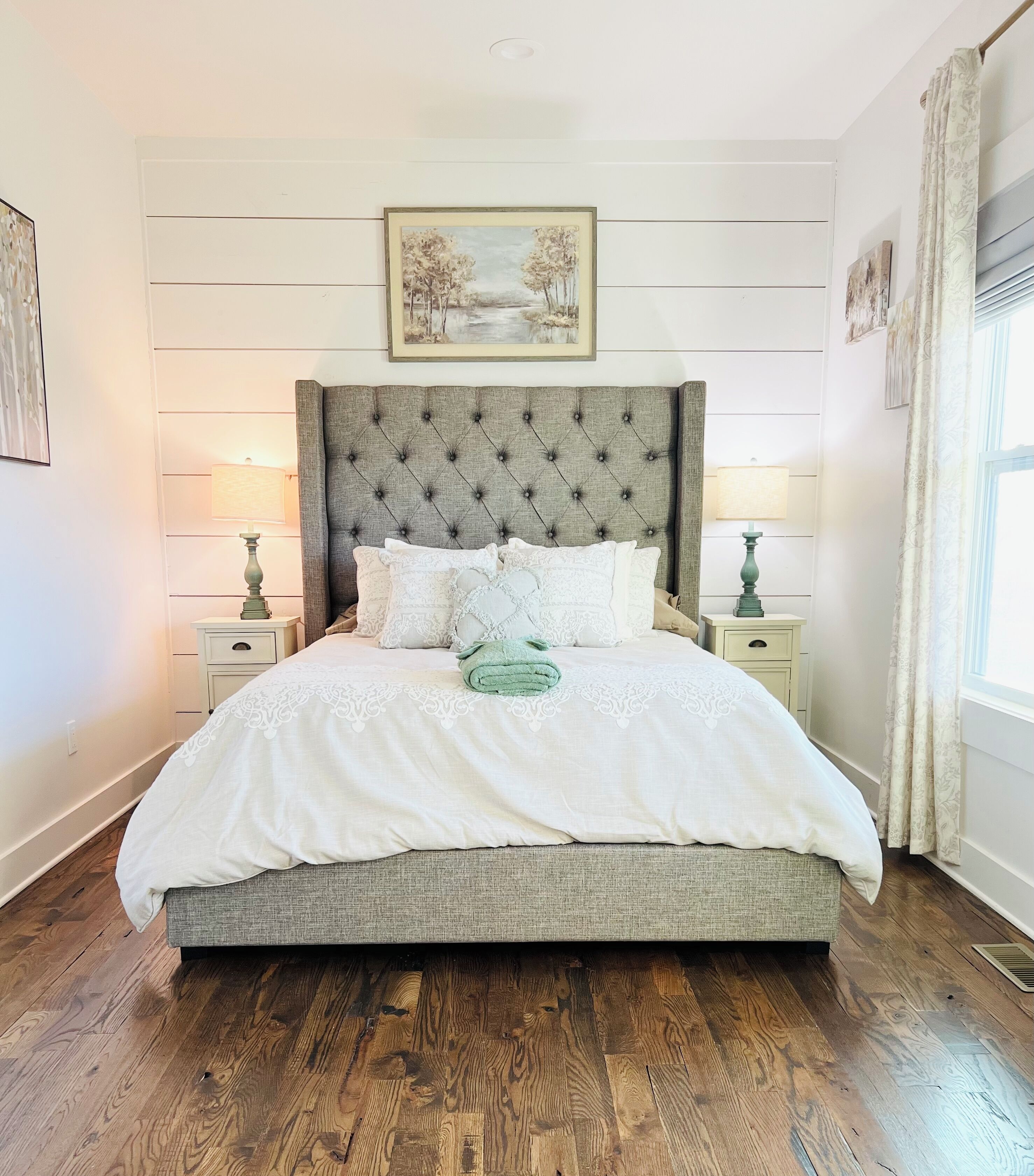 A neatly made bed with a tufted headboard, two bedside tables with lamps, a framed landscape painting above the bed, and a window with curtains.