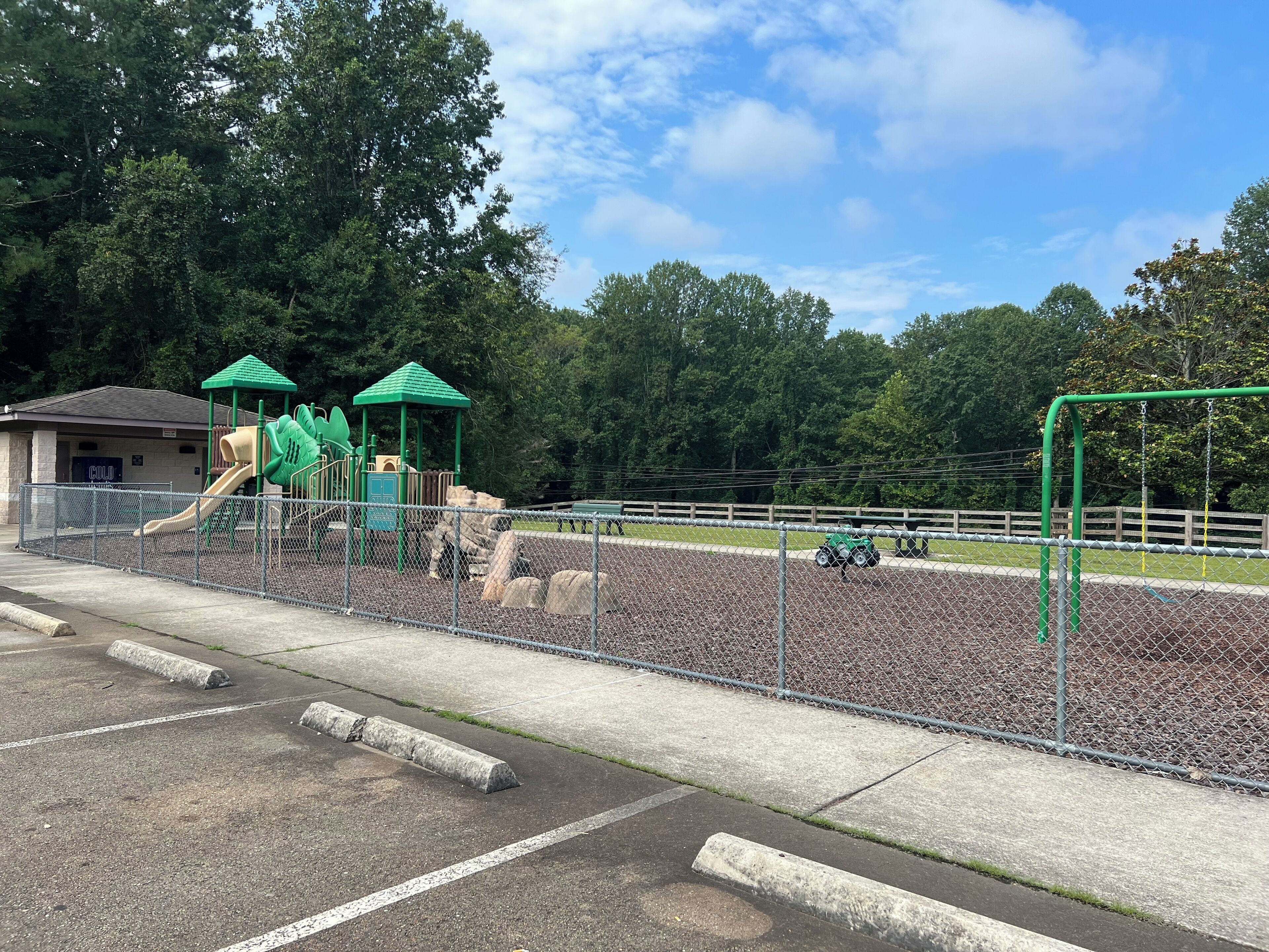 A playground with a slide and climbing structures, surrounded by a fence and trees.
