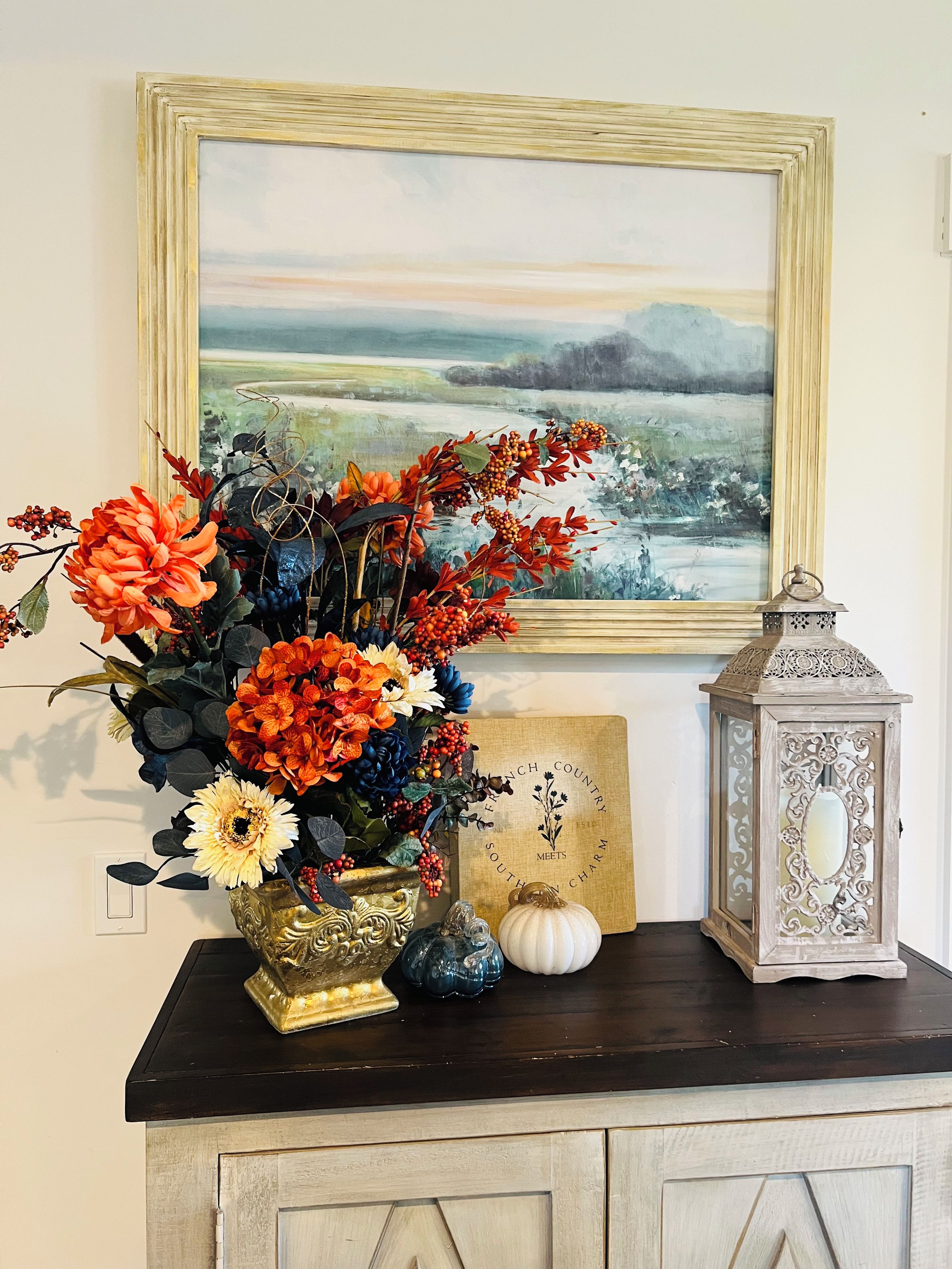 A decorative arrangement on a dark wooden surface including a vase with flowers, a lantern, and a framed picture.