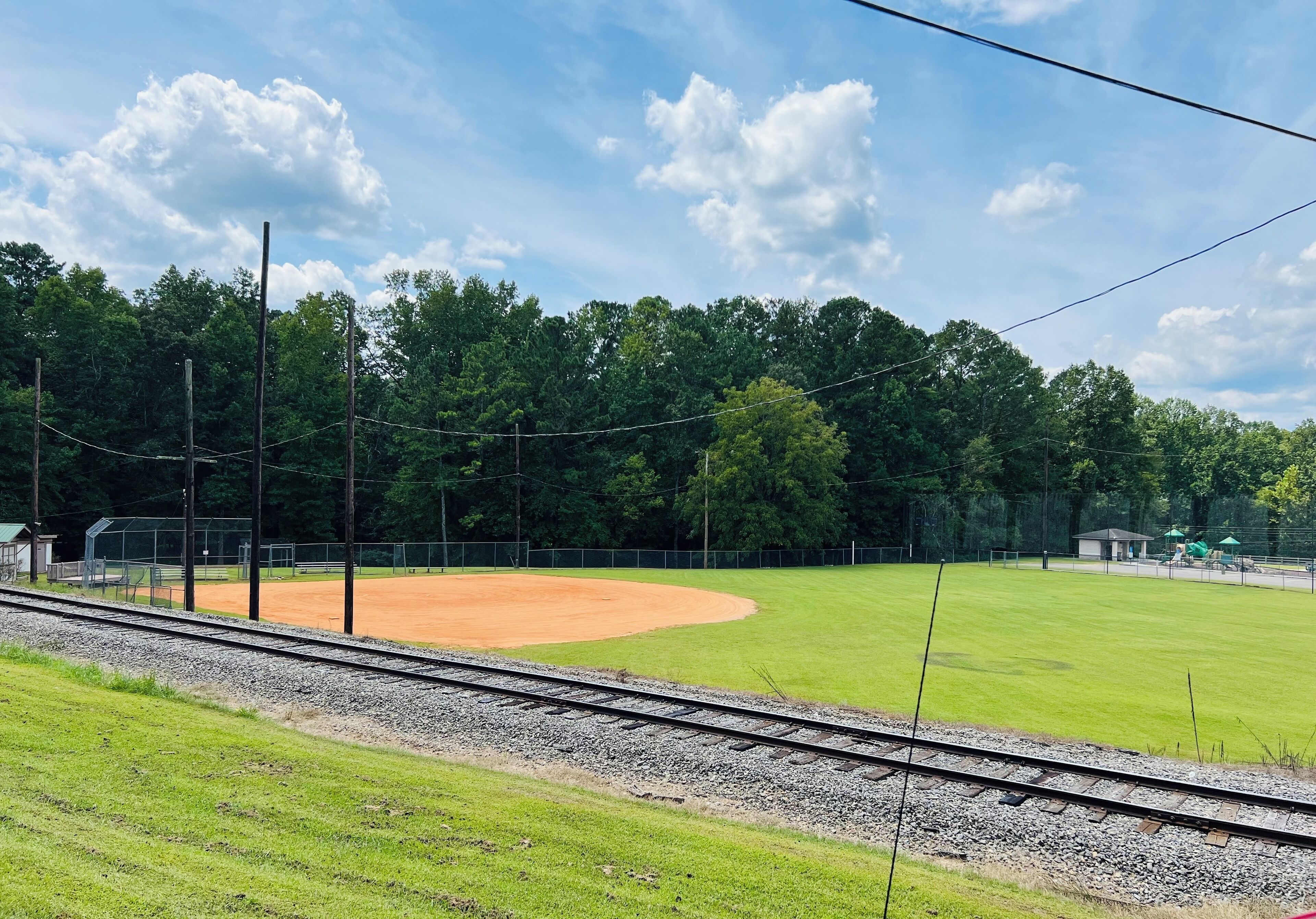 Ein Baseballfeld mit erdschläglichem Innenfeld und grünem Außenfeld, umgeben von Bäumen und einem Zaun.