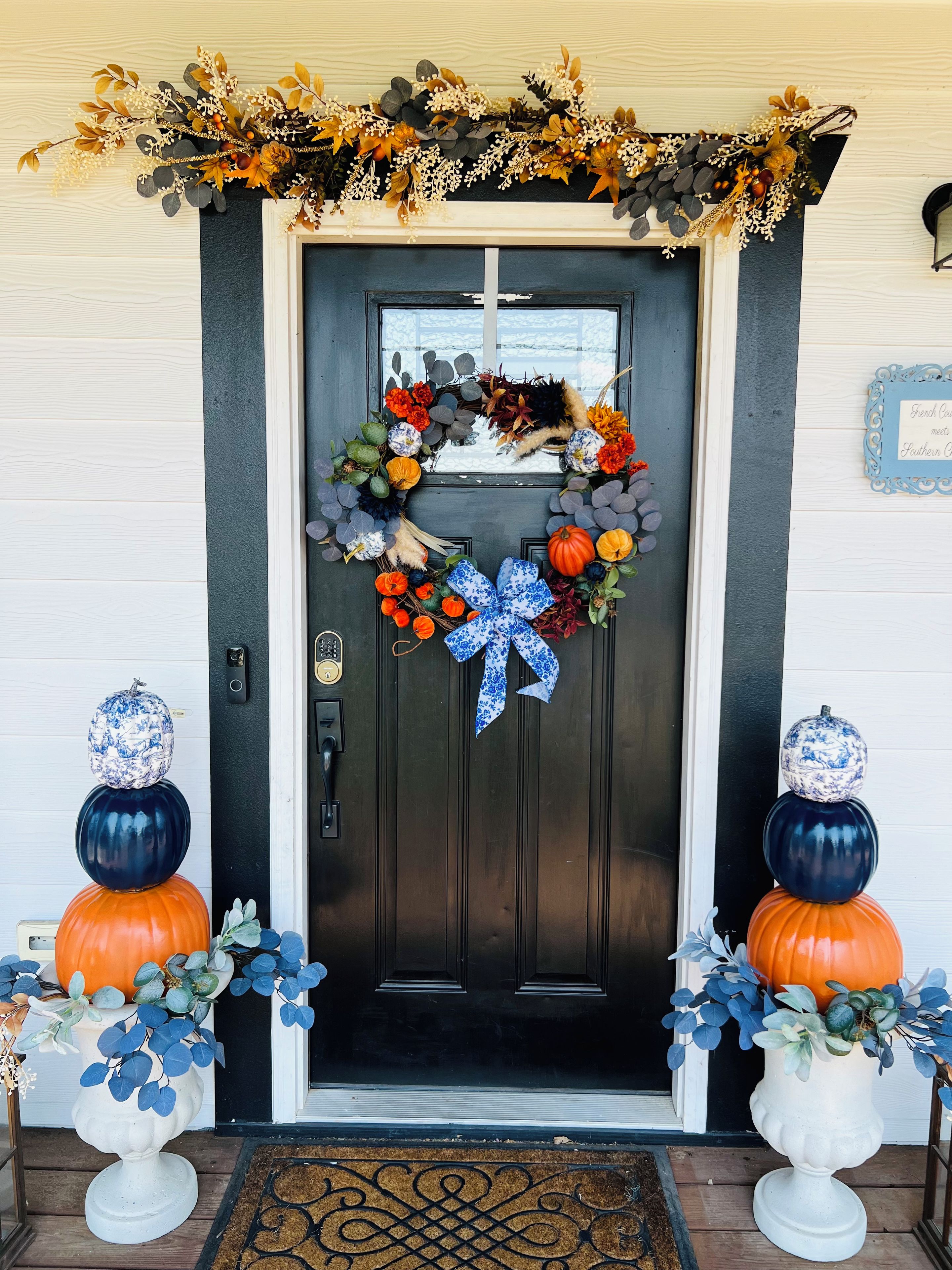 Una puerta principal con un adorno de lazo, dos calabazas decorativas y un felpudo.