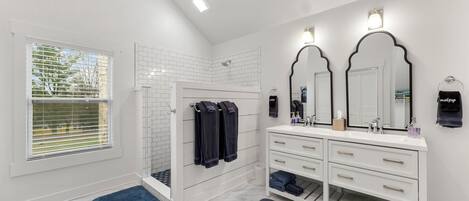 A modern bathroom with white tiled walls, a walk-in shower, a vanity with two mirrors, and a window with blinds.