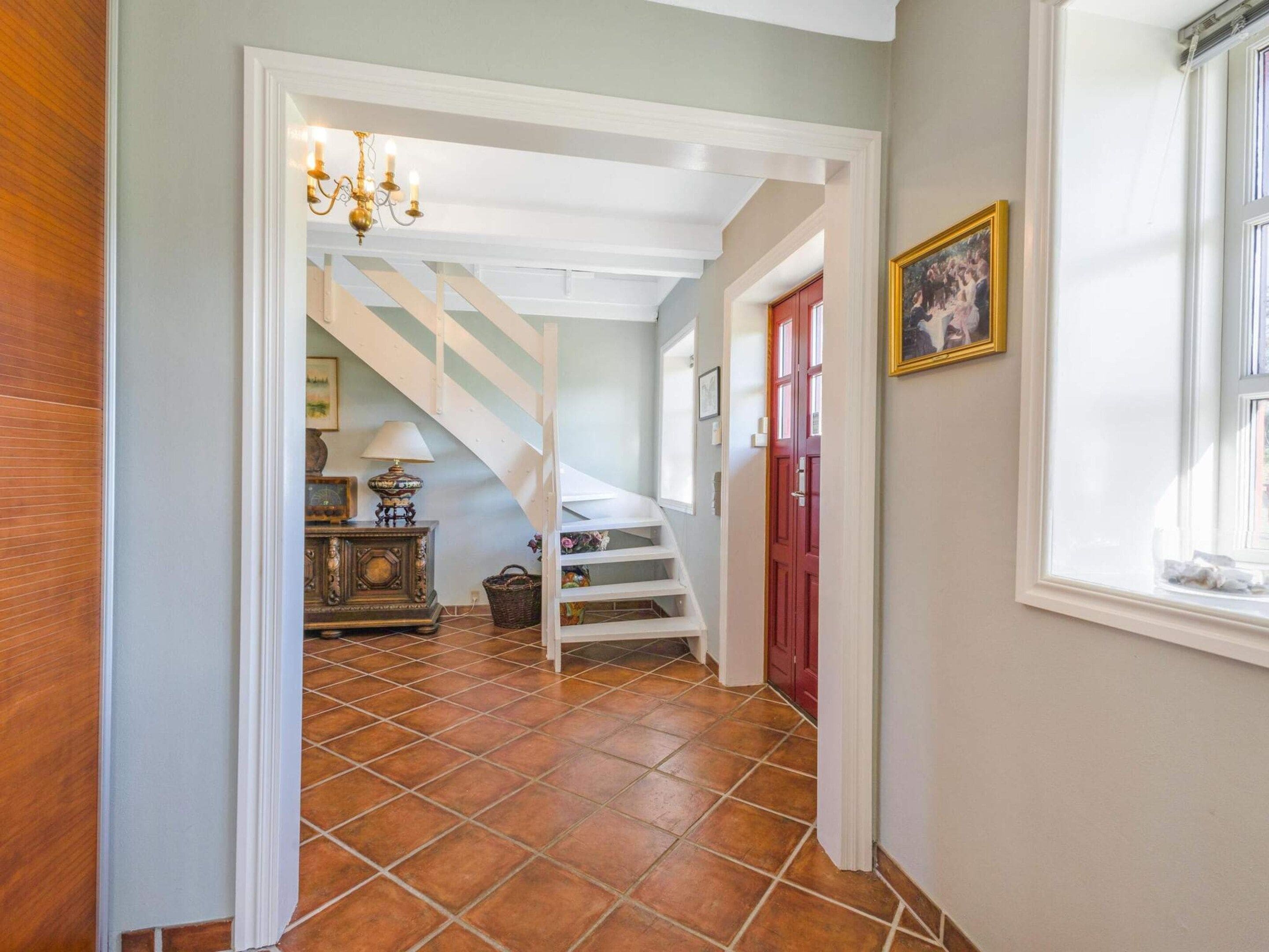Un pasillo con una escalera, un candelabro, una puerta con un cuadro y una ventana.