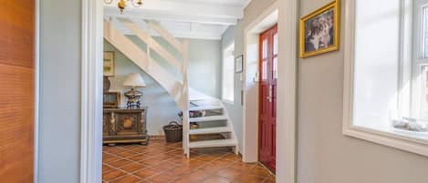 A hallway with a staircase, a chandelier, a door with a picture, and a window.