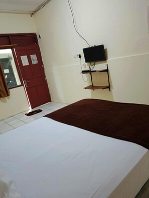 Una habitación con cama, televisor en la pared, una puerta roja y una ventana con cortinas.