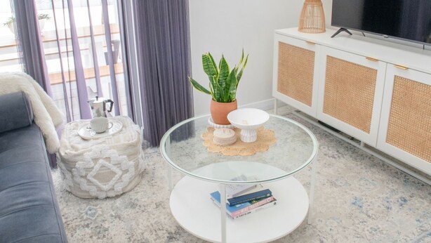 A modern living room with a sofa, a glass coffee table with books, a potted plant, a TV, and a cabinet with a woven front panel. - Welcome to Bay Belle - Binalup (MIDDLETON BEACH)