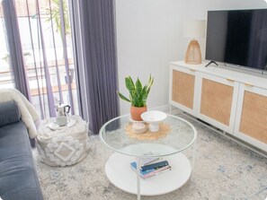 A modern living room with a sofa, a glass coffee table with books, a potted plant, a TV, and a cabinet with a woven front panel.