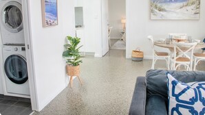 A modern living room with a grey sofa, a dining area with a round table and chairs, a kitchen with a washing machine, and a hallway with a plant and artwork. - Welcome to Bay Belle - Binalup (MIDDLETON BEACH)