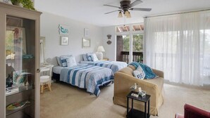 A bedroom with two beds, a ceiling fan, a window with sheer curtains, and a view of greenery outside.