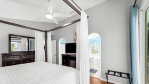 Ein Schlafzimmer mit einem großen Bett, einem Deckenventilator, einem Fernseher und Blick auf einen Balkon mit blauem Himmel.