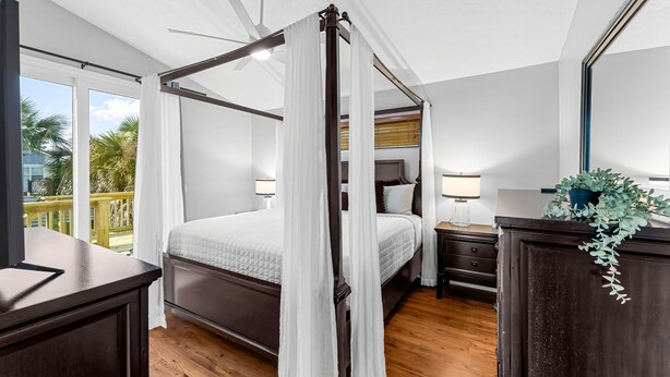 Ein Bett mit Baldachin, ein Deckenventilator, ein Nachttisch mit Lampe und Blick ins Freie durch große Fenster.