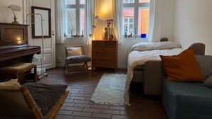 A cozy room with a piano, a sofa, a chair, and a bed. There are two windows with curtains, a dresser, and a lamp.