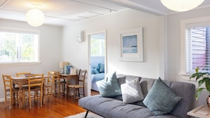 En stue med en grå sofa, et træspisebord og stole. Billeder hænger på væggene, og et vindue har persienner.