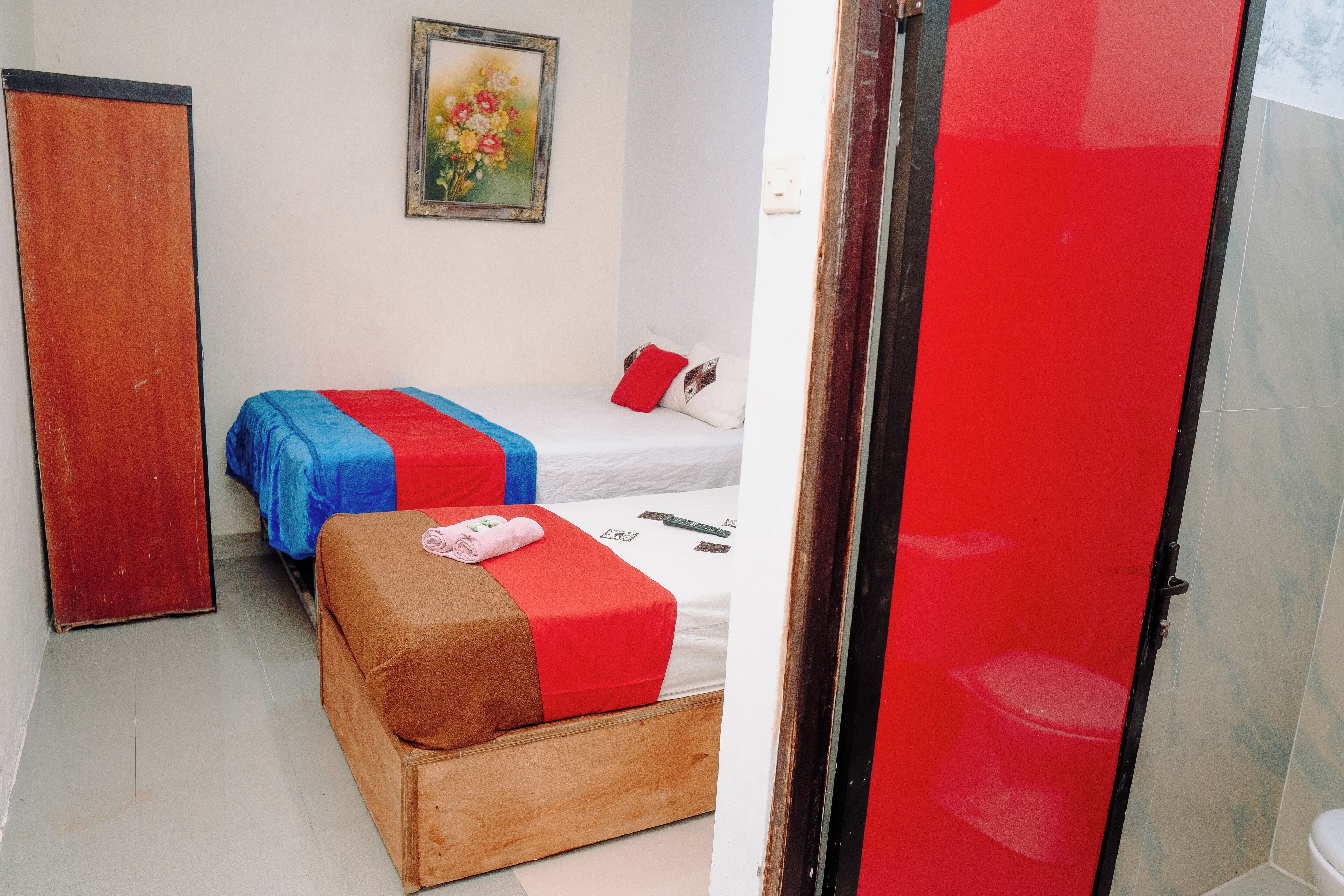 Une pièce avec deux lits, un banc en bois, une porte rouge et un tableau accroché au mur.