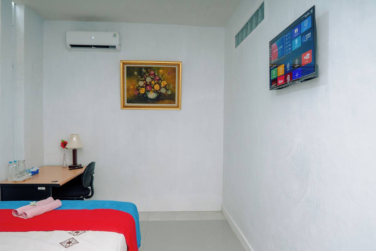 Une chambre d’hôtel comprenant un lit, un bureau avec une lampe, un tableau, un climatiseur et un écran numérique.