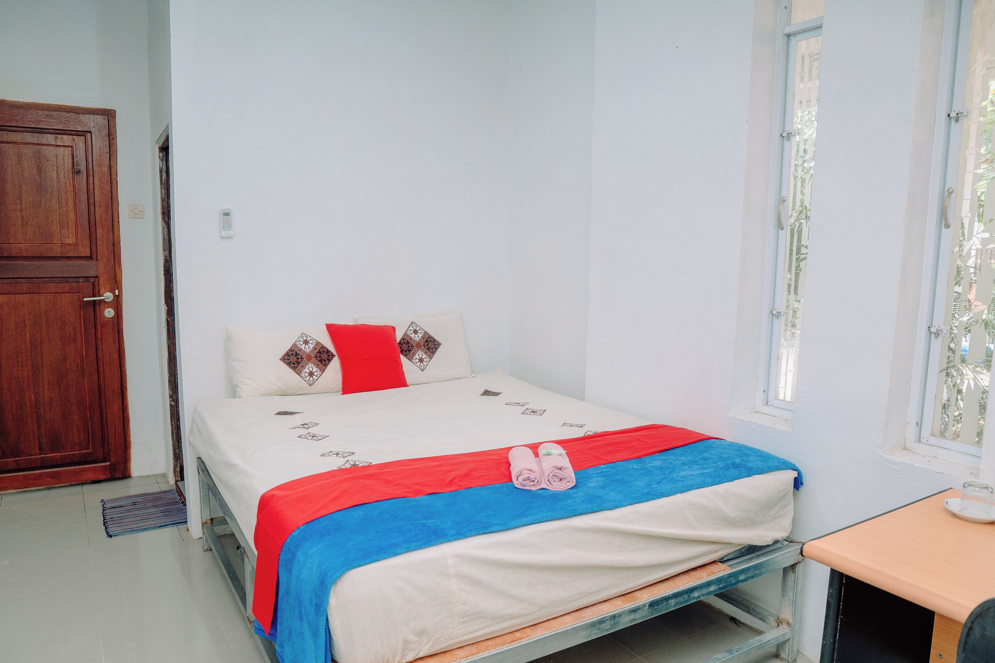 Un lit simple avec une couverture rouge et bleue, une porte en bois et une fenêtre donnant sur un paysage verdoyant.