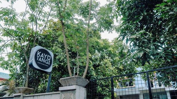 Señalización para 'KAYO TANGI GUEST HOUSE SYARIAH' con un muro de concreto y una valla metálica al frente.