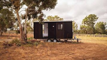 A small, dark-colored mobile home with a sliding door, situated in a dry, grassy area with trees in the background.