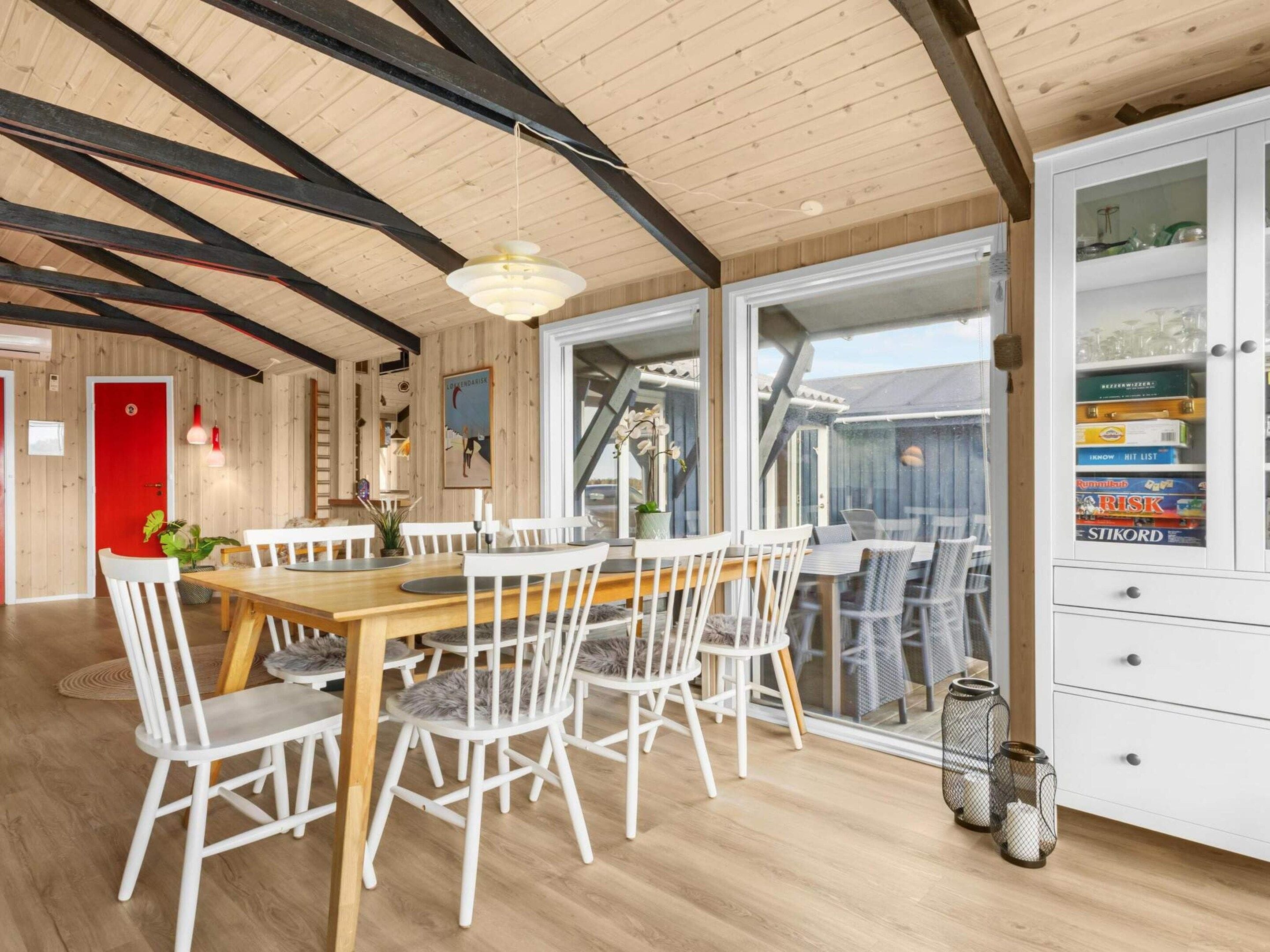 A dining area with a wooden table and white chairs, a red door, and a cabinet with board games.