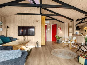 A cozy interior with a wooden ceiling, a flat-screen TV mounted on the wall, a dining area with a wooden table and chairs, and a red door leading to another room. - 5 Star Holiday Home in Lokken (Lokken)