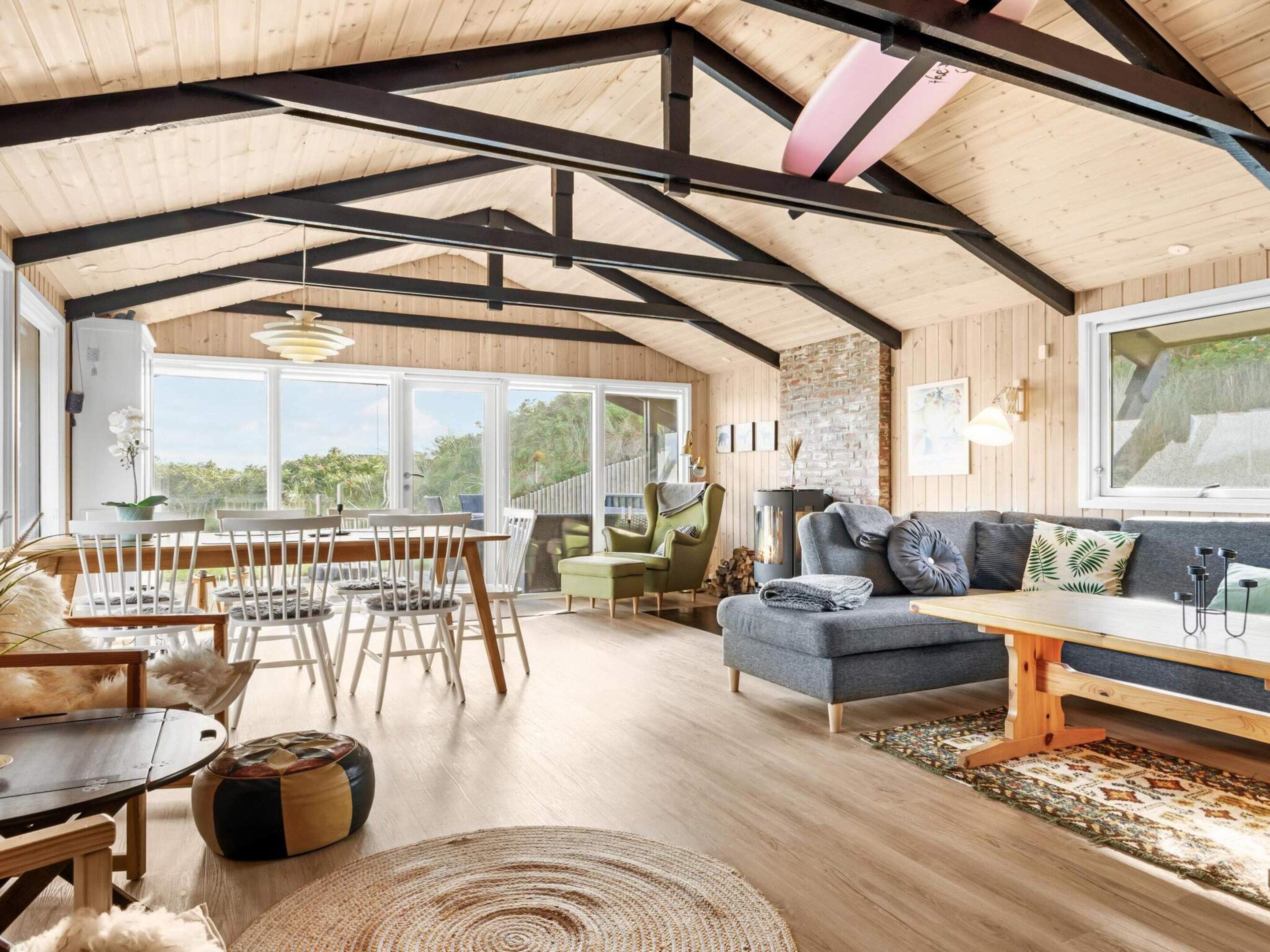 A spacious living area with a high ceiling, wooden beams, a sofa, a dining table with chairs, and a large window offering a view of greenery.